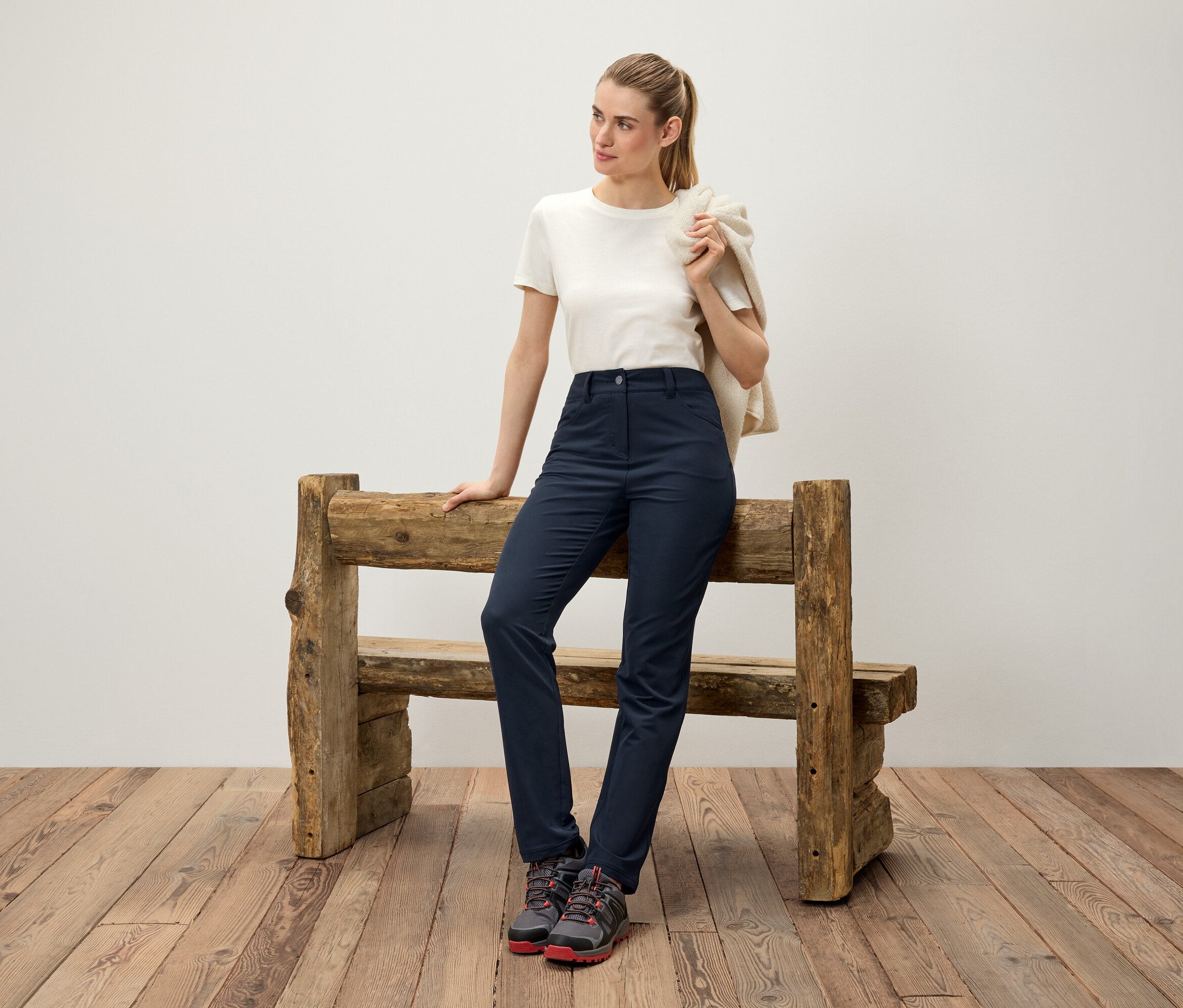 Eine Frau lehnt an einer Holzbank, trägt einen cremefarbenen Pullover über der Schulter, ein weißes T-Shirt, dunkelblaue Hosen und Trekkingschuhe.