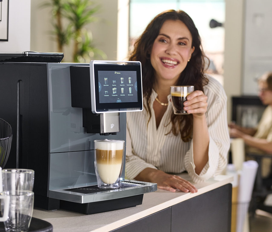 Frau hält ein Glas Kaffee neben einer Kaffeemaschine.