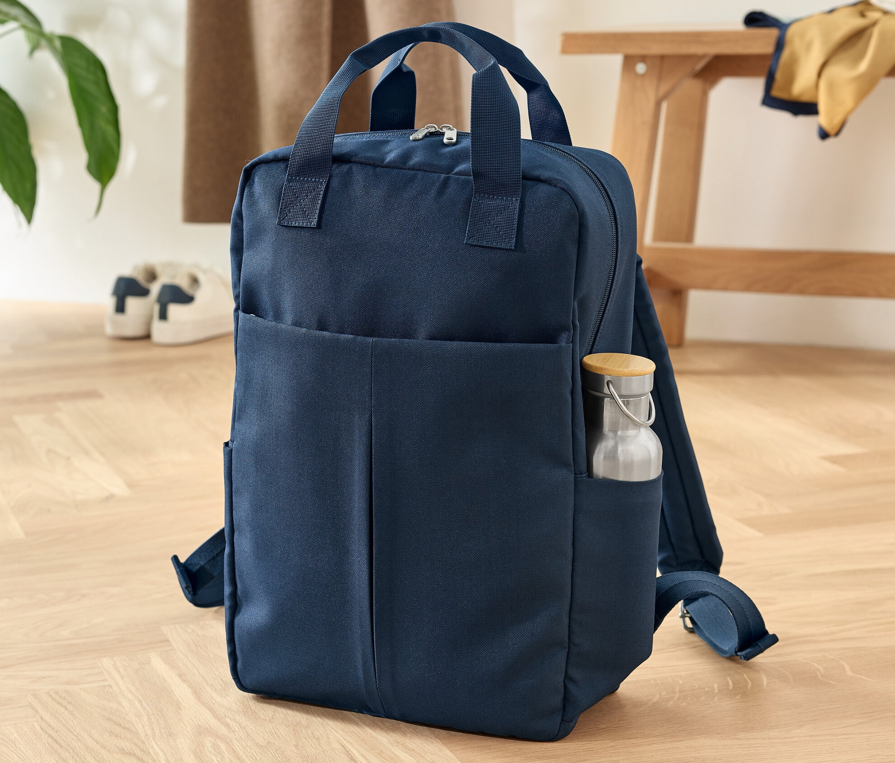 Ein marineblauer Sicherheits-Rucksack steht auf einem Holzboden. In der Seitentasche befindet sich eine Wasserflasche.