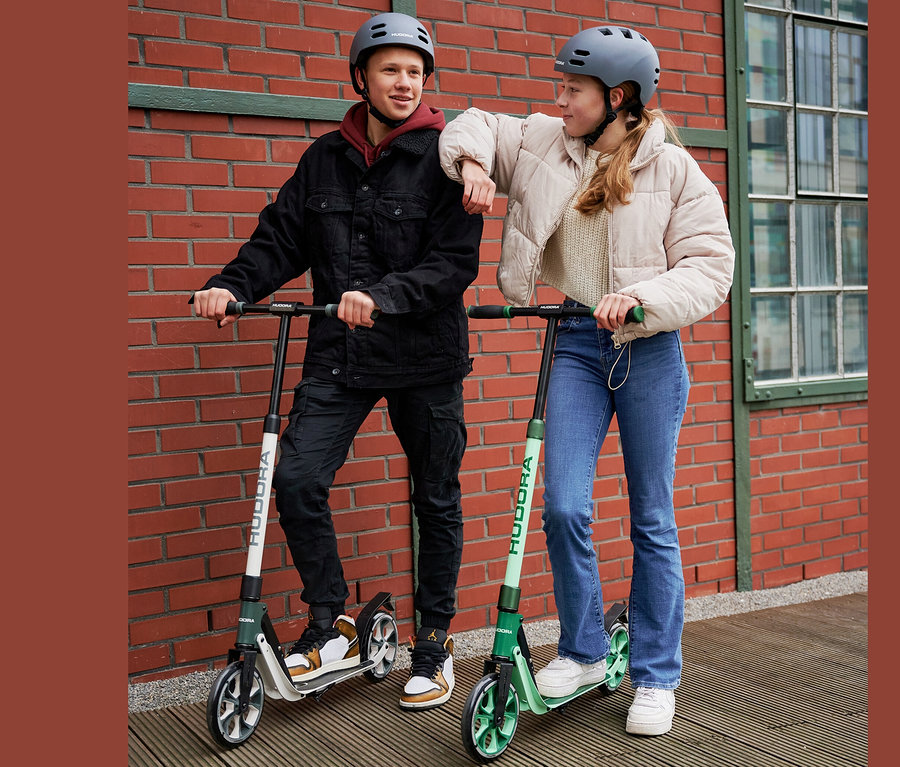 Ein junger Mann und eine junge Frau mit grauen Helmen stehen an einer Ziegelmauer auf HUDORA BigWheel® 205 Rollern, einer grau-weiß, der andere grün.