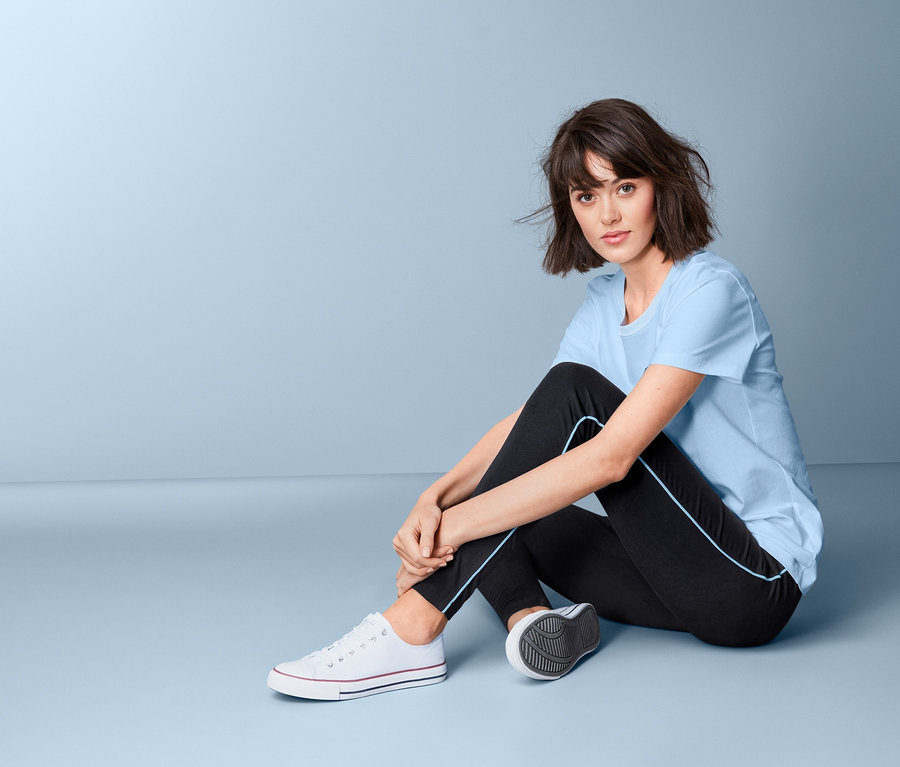 Frau sitzt in Leggings mit Paspelierung und hellblauem T-Shirt auf dem Boden.