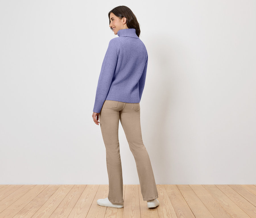 Frau steht mit dem Rücken zur Kamera, trägt einen lila Rollkragenpullover und beige Hosen.