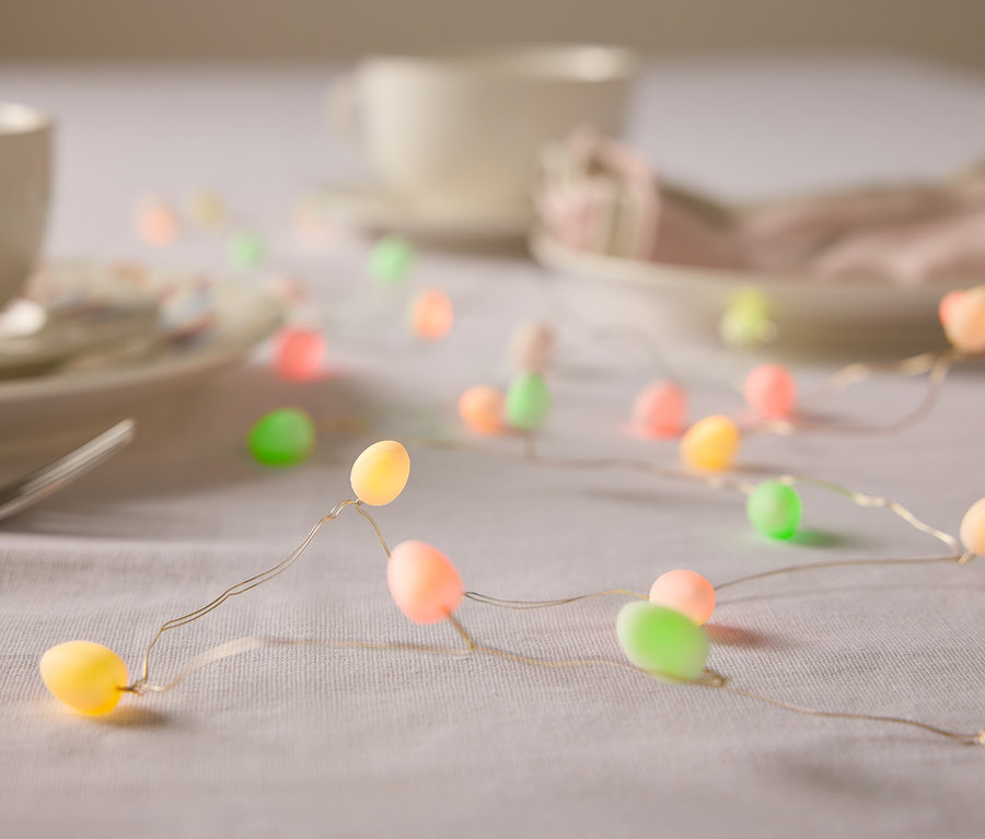 Nahaufnahme von einem Tisch mit Geschirr und einer Lichterkette in Form von Ostereiern in Pastellfarben.