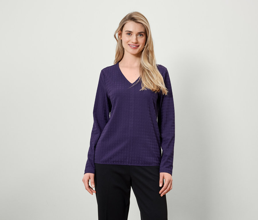 Blonde Frau mit langen Haaren posiert in einer violetten Langarmbluse und einer schwarzen Hose.