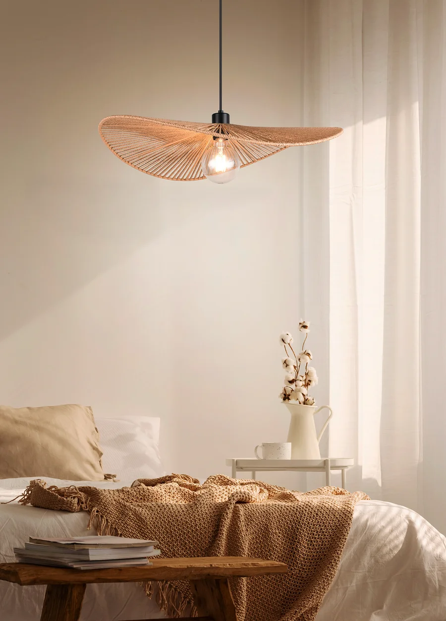Eine strohfarbene Lampen-Hängelampe in Hutform hängt über einem Bett in einem Schlafzimmer.