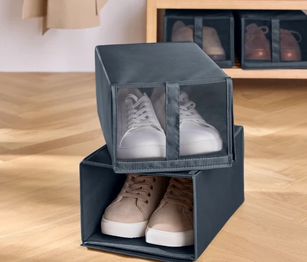 Zwei graue Stoff-Schuhboxen sind übereinander gestapelt. In der oberen Box befinden sich weiße Turnschuhe und in der unteren Box braune Turnschuhe. Im Hintergrund befindet sich ein Holzregal mit Schuhen.