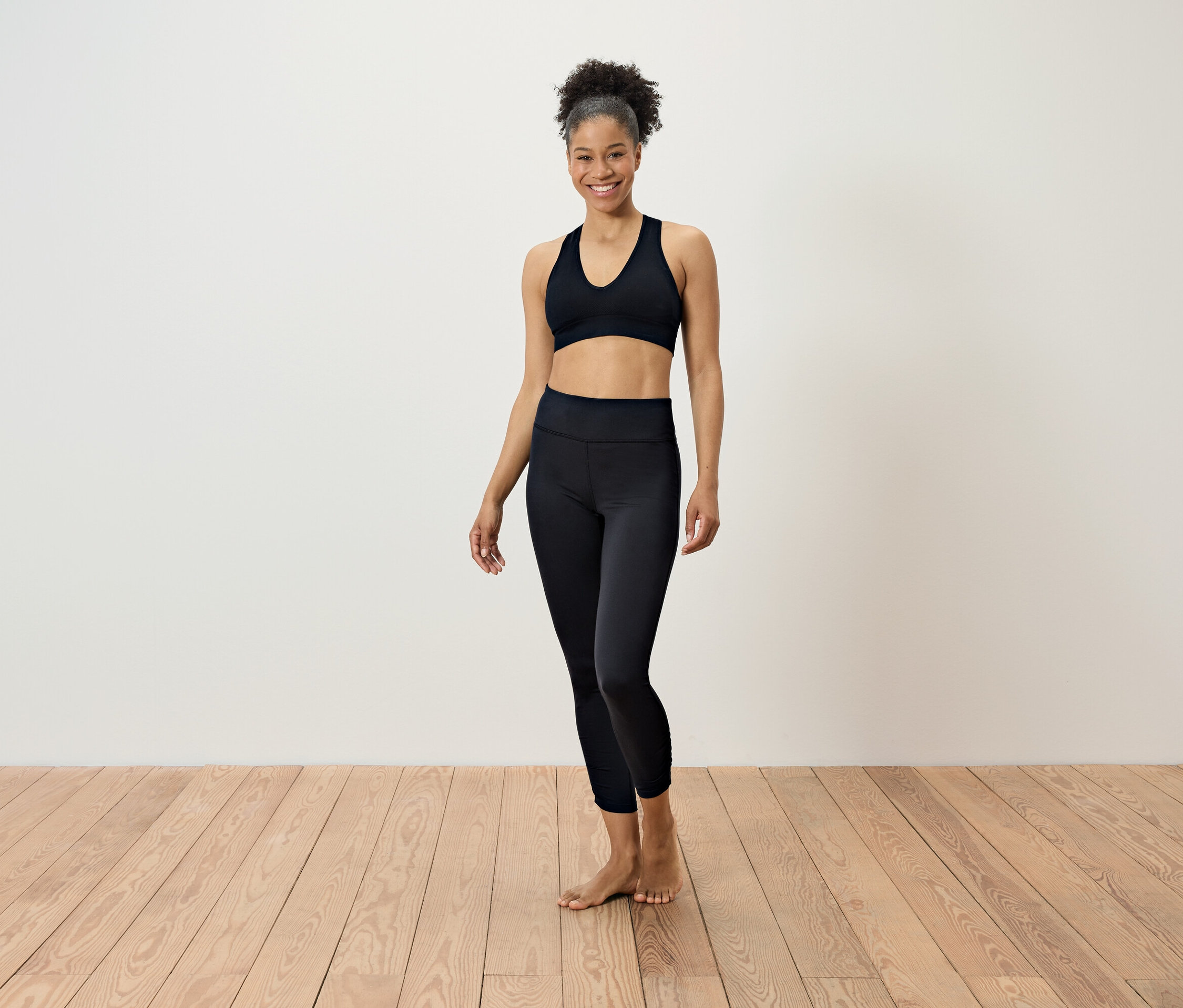 Frau in schwarzer Sportbekleidung steht auf einem Holzboden vor einer weißen Wand.