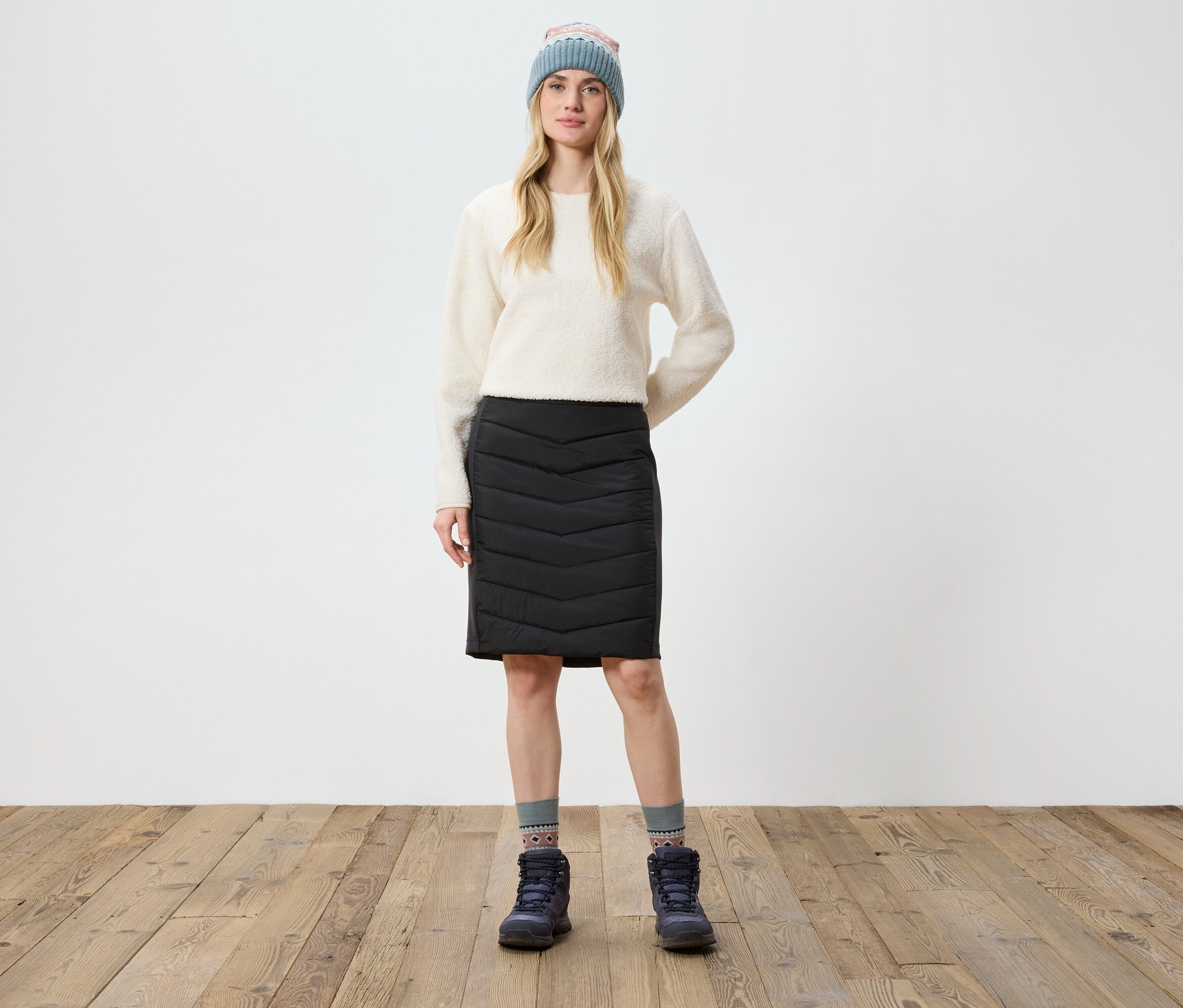 Eine Frau steht auf einem Holzboden und trägt einen cremefarbenen Pullover, einen schwarzen Rock, Stiefel und eine Strickmütze.