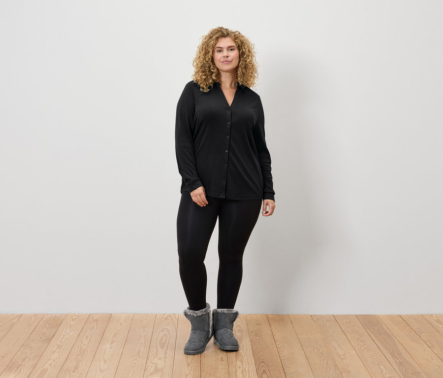 Eine Frau mit blonden Haaren steht und trägt schwarze Leggings, ein schwarzes Langarmhemd und graue Stiefel.