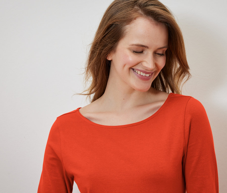 Lächelnde Frau mit langen Haaren in einem orangefarbenen Langarmshirt.