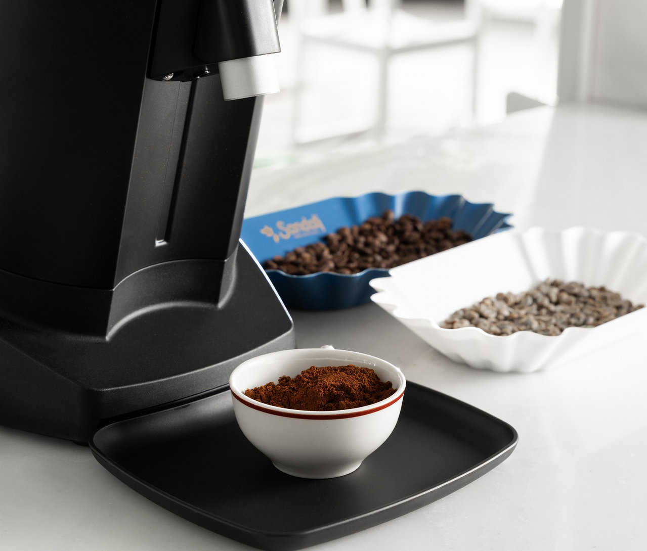 Kaffeemühle neben einer Schale mit Kaffeepulver, einer blauen Schale mit Kaffeebohnen und einer weißen Schale mit helleren Kaffeebohnen.