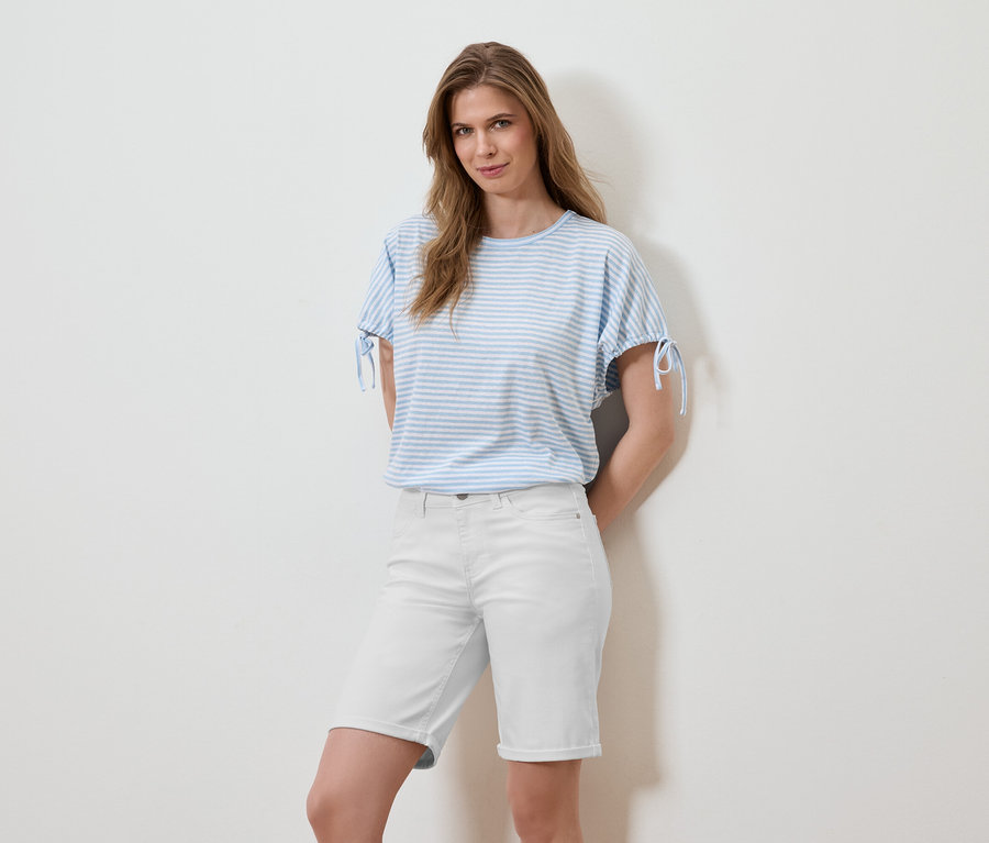 Frau posiert in einem gestreiften, hellblauen T-Shirt und weißen Shorts vor einer weißen Wand.