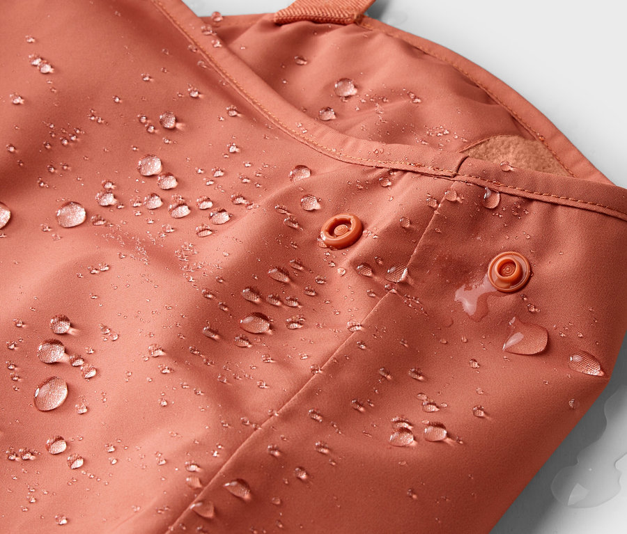 Nahaufnahme einer wasserabweisenden, terrakottafarbenen Jacke mit Druckknöpfen und Wassertropfen.