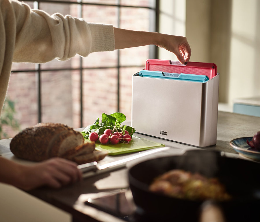Eine Hand nimmt ein pinkes Schneidebrett aus einem Behälter. Auf dem Tisch liegen Brot, Radieschen und eine Pfanne.