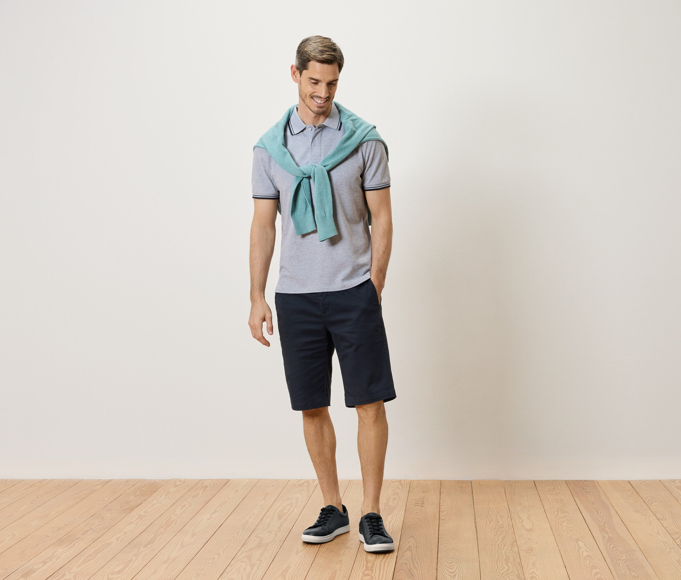 Mann steht mit einem hellblauen Pullover um den Hals, einem grauen Poloshirt, einer dunkelblauen Shorts und blauen Turnschuhen.