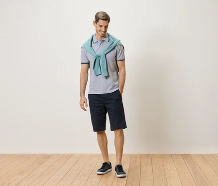 Mann steht mit einem hellblauen Pullover um den Hals, einem grauen Poloshirt, einer dunkelblauen Shorts und blauen Turnschuhen.