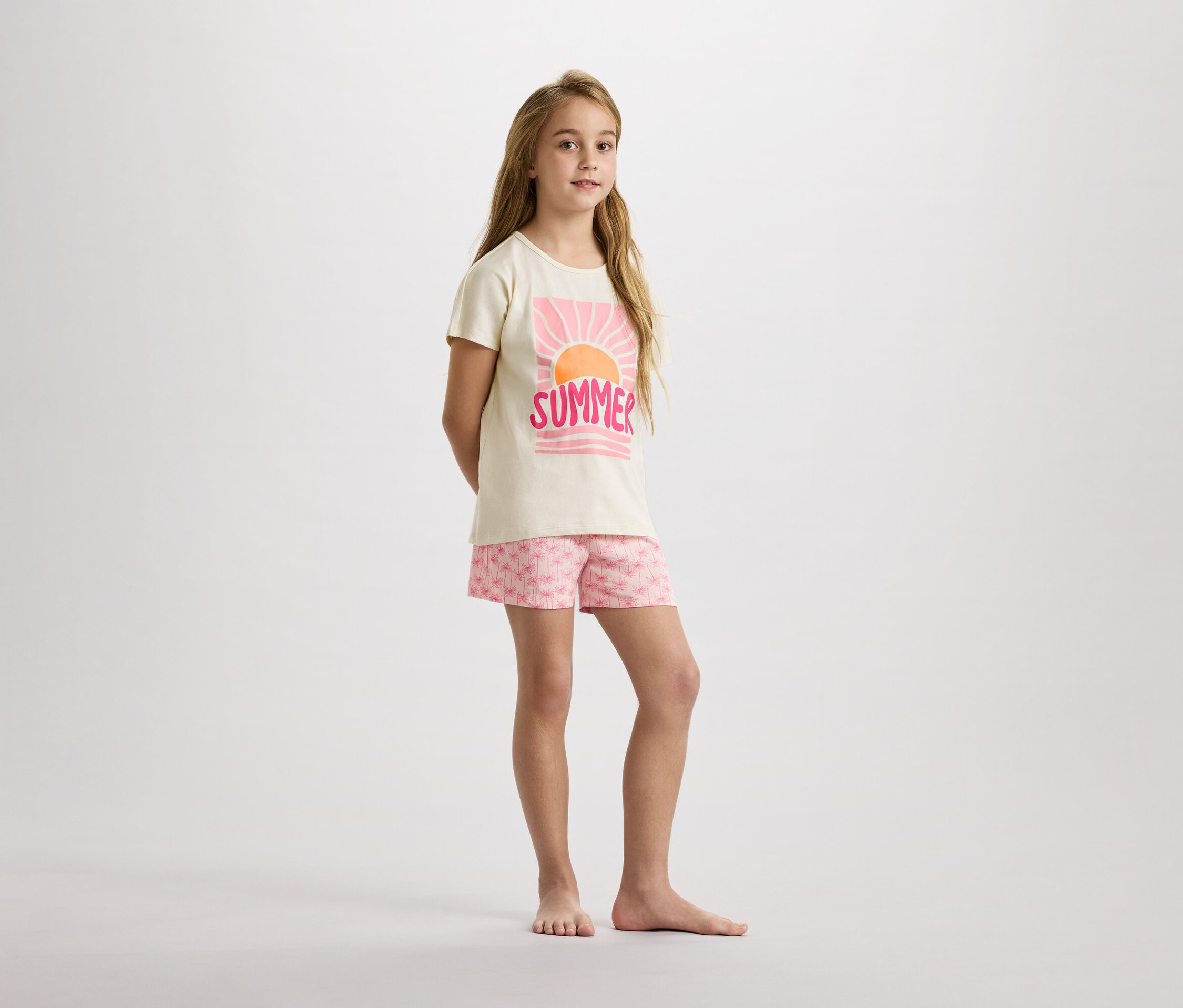 Ein junges Mädchen steht barfuß in einem beigen T-Shirt mit einem rosa "Summer"-Aufdruck und Shorts mit rosa Palmenmuster vor einem weißen Hintergrund.