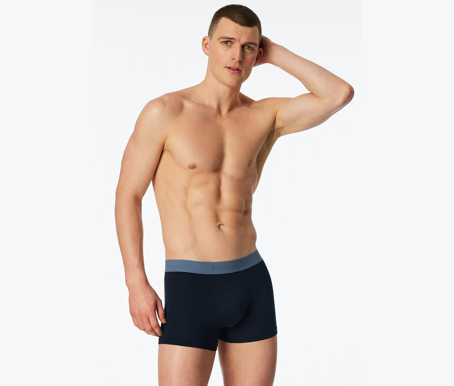 Mann ohne Hemd posiert in marineblauer Boxershorts mit blauem Bund vor einem weißen Hintergrund.