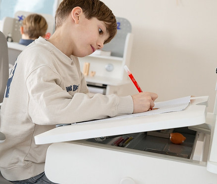 Ein Junge sitzt an einem weißen Schreibtisch und schreibt mit einem roten Stift auf ein Blatt Papier. Eine Schublade ist geöffnet.