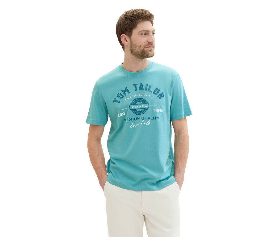 Mann mit Tom Tailor T-Shirt mit Logo in Meadow Teal und beigen Hosen, die Hände in den Hosentaschen.