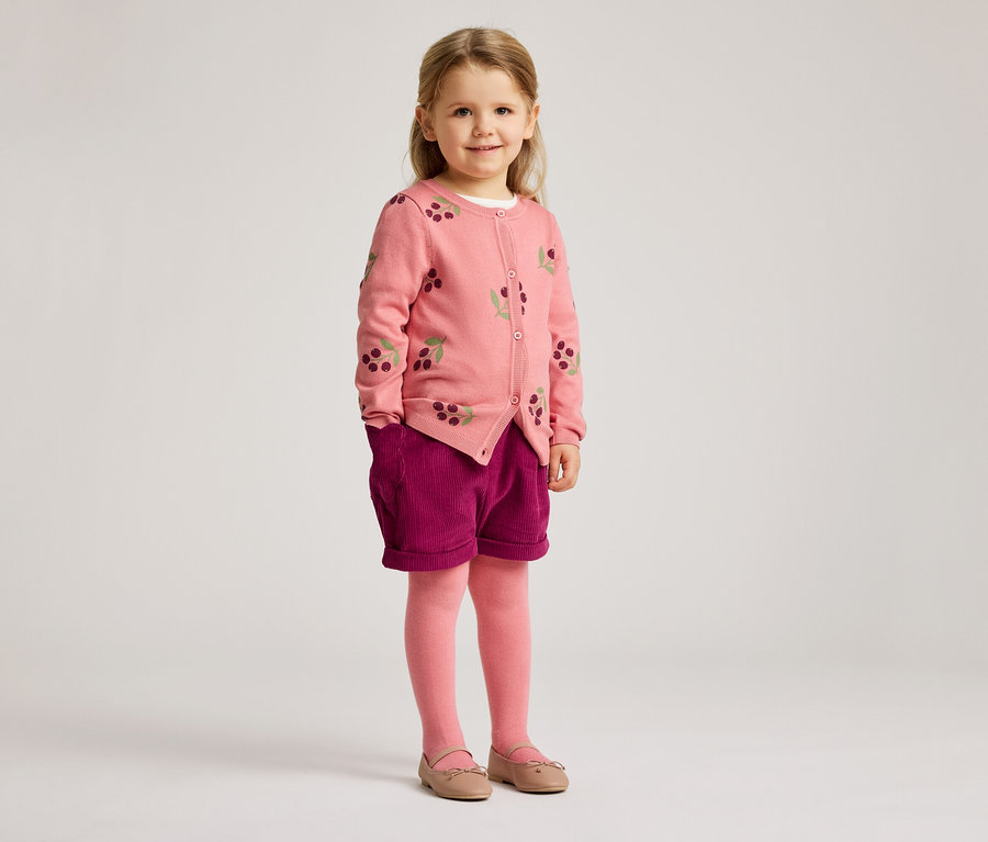 Ein kleines Mädchen mit blonden Haaren trägt eine rosa Strickjacke mit Kirschmuster, violette Shorts, rosa Strumpfhosen und Ballerinas.