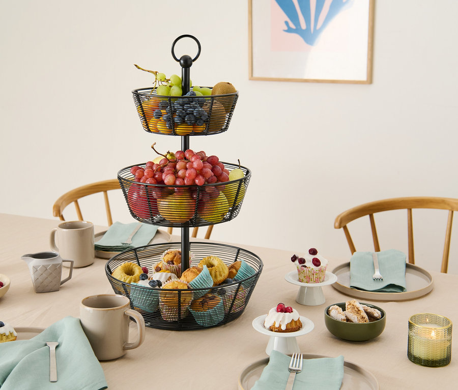 Ein drei stöckiger Obstkorb aus Metalldraht steht auf einem Tisch mit Gebäck, Obst und Geschirr.
