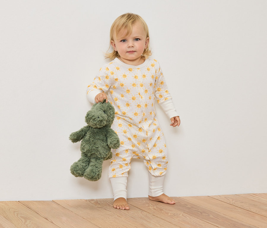 Ein blondes Kind im Overall mit Sonnenmuster hält einen grünen Stoffdinosaurier.
