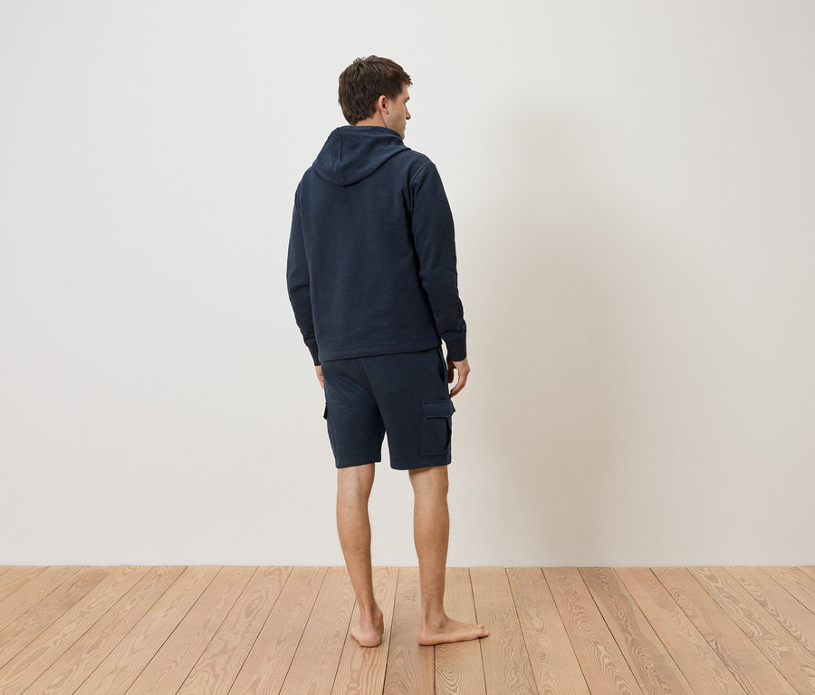 Mann von hinten, trägt einen dunkelblauen Hoodie und Shorts auf einem Holzboden.