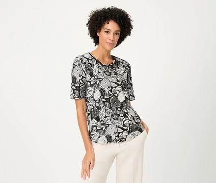 Frau mit lockigem Haar trägt ein Olsen Shirt mit Blumenmuster und helle Hosen.