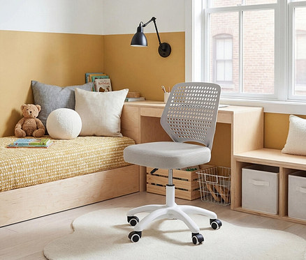 Ein Kinderzimmer mit einem Bettsofa, einem Schreibtisch mit Lampe und einem grauen Bürostuhl auf einem weißen Teppich.