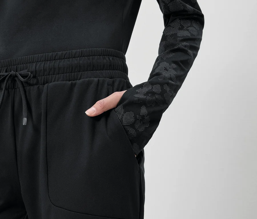 Frau mit Hand in der Tasche einer schwarzen Jogginghose mit Kordelzug und einem schwarzen, langärmeligen Oberteil mit Muster.