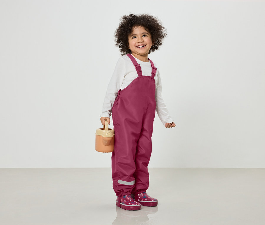 Ein lächelndes Kind mit lockigem Haar trägt eine weinrote Latzhose und Gummistiefel mit Blumenmuster und hält eine hellbraune Gießkanne.