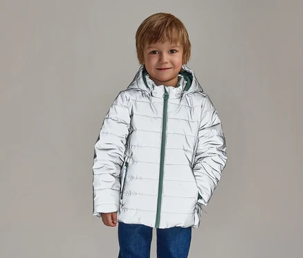 Ein lächelnder Junge mit blonden Haaren trägt eine reflektierende, silberne Pufferjacke mit Kapuze und grünem Reißverschluss über blauen Jeans.