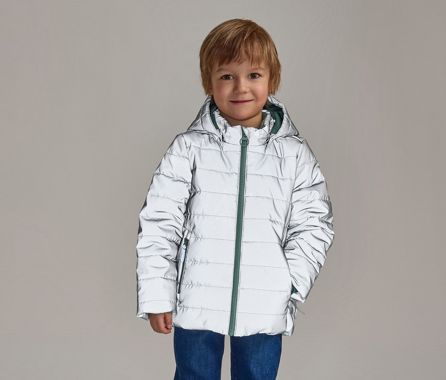 Ein lächelnder Junge mit blonden Haaren trägt eine reflektierende, silberne Pufferjacke mit Kapuze und grünem Reißverschluss über blauen Jeans.