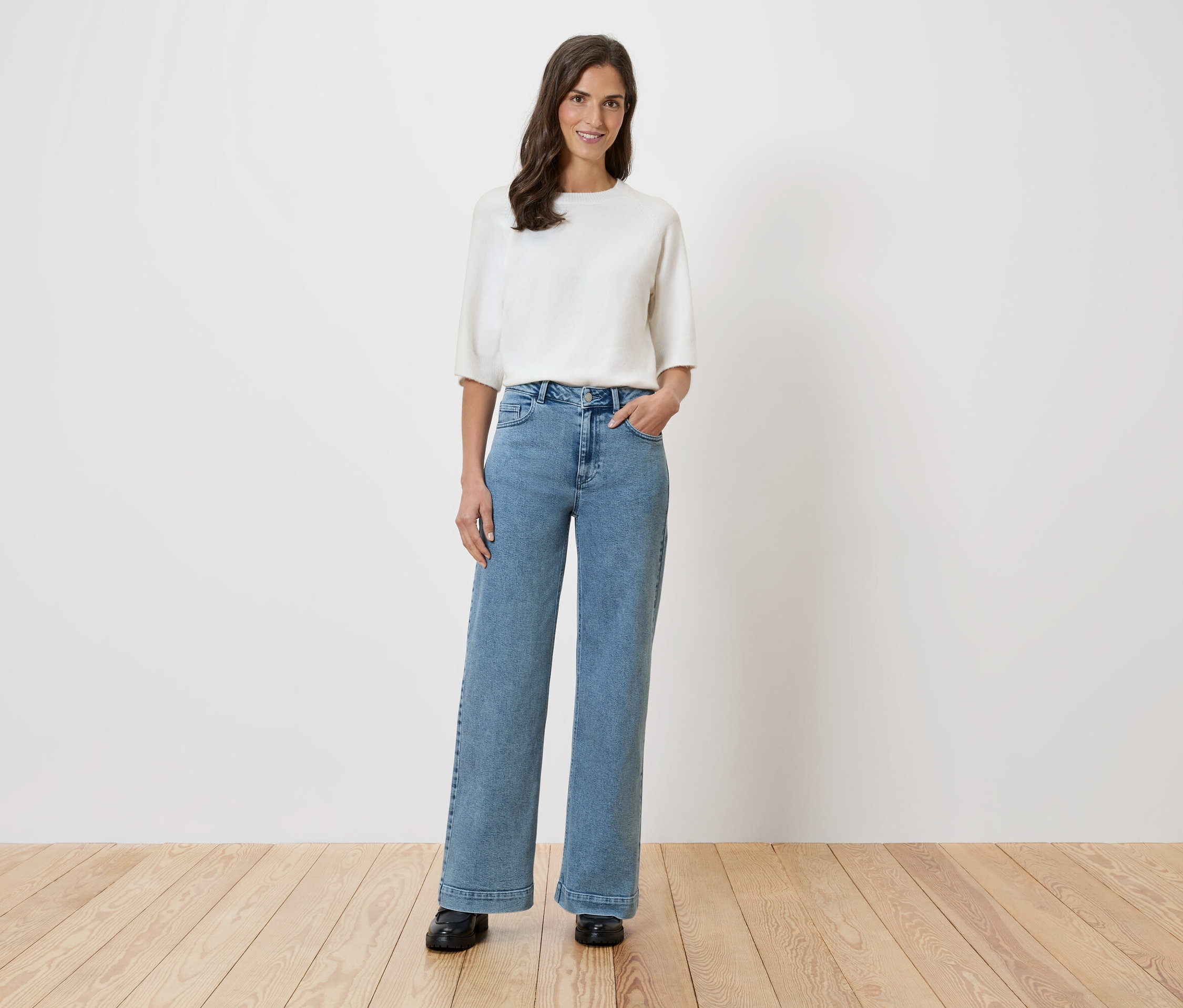 Eine Frau steht vor einer weißen Wand und trägt eine weiße Bluse und weite Jeans.