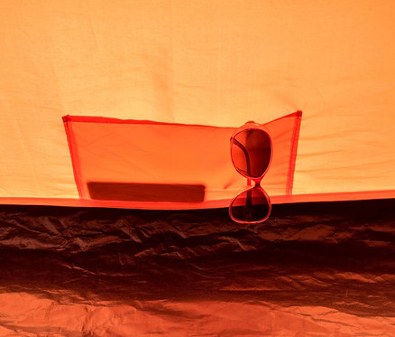 Innenansicht eines orangefarbenen Tambu »HAIKA 2« 2-Personen-Kuppelzelts mit aufgesetzter Tasche und einer Sonnenbrille.