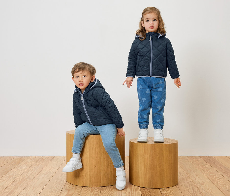 Ein Junge sitzt auf einem Holzhocker, während ein Mädchen auf einem anderen steht. Beide tragen marineblaue, gesteppte Jacken und Jeans.