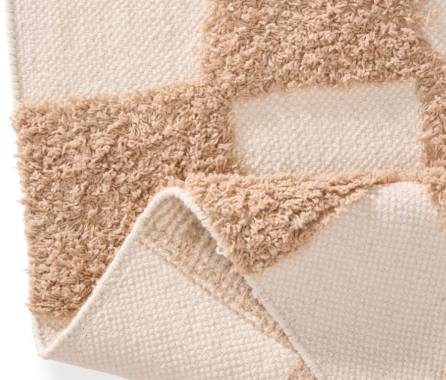 Nahaufnahme eines beige-farbenen Teppichs mit Schachbrettmuster, dessen Ecke umgeschlagen ist.