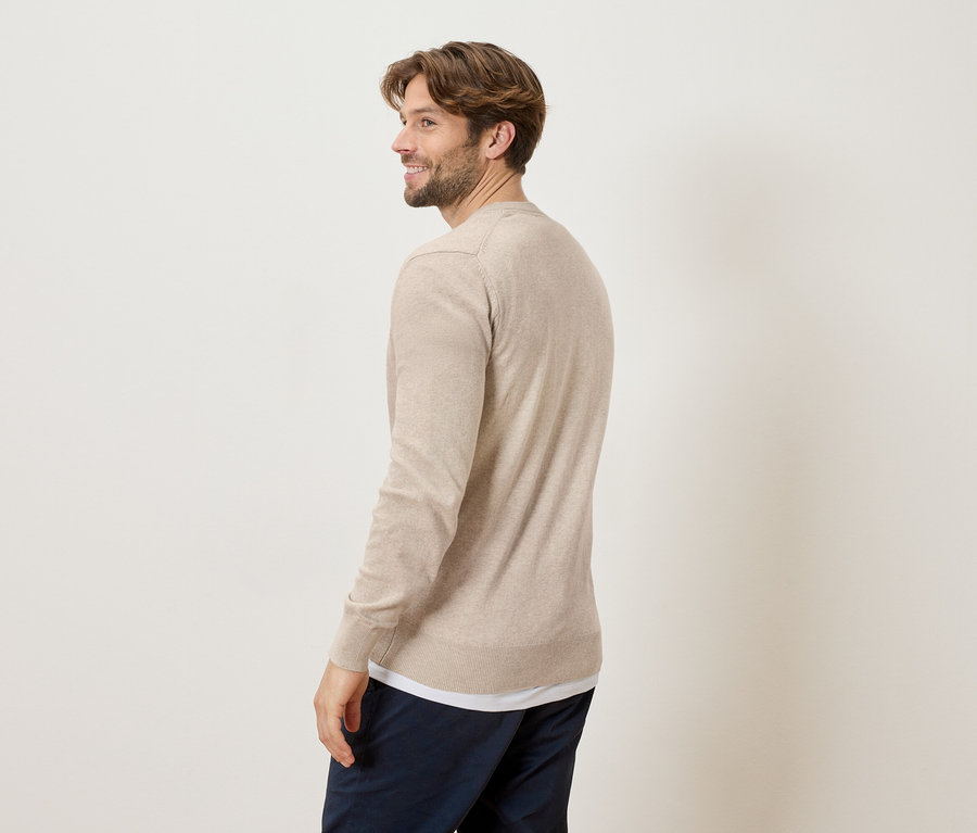 Ein Mann blickt über seine Schulter und trägt einen beige Pullover und marineblaue Hosen vor einem cremefarbenen Hintergrund.