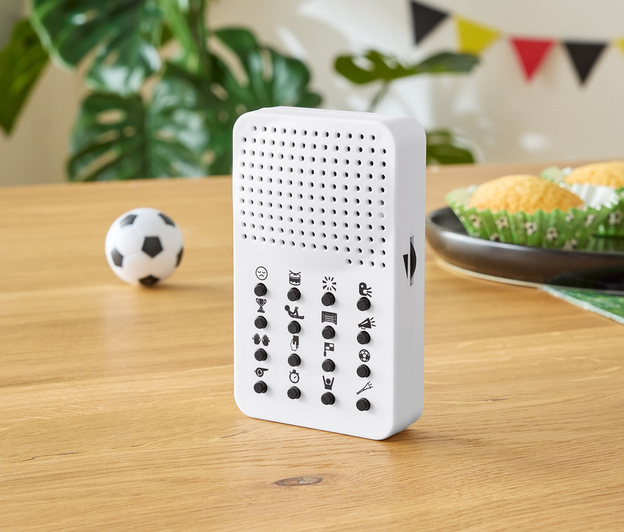Weißes Gerät mit Knöpfen auf einem Holztisch, mit einem kleinen Fußball, Muffins und Partydekorationen im Hintergrund.