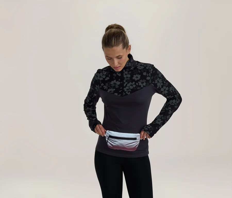 Frau in einem dunkelgrauen Langarmshirt mit schwarzem Blumenmuster an Schultern und Ärmeln, schwarzen Leggings und einer silbernen Bauchtasche, die auf die Bauchtasche blickt.