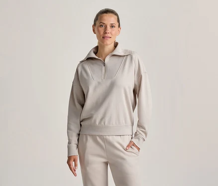 Frau steht, trägt ein hellbeiges Half-Zip-Sweatshirt und passende Jogginghose, eine Hand in der Tasche, blickt nach vorne.