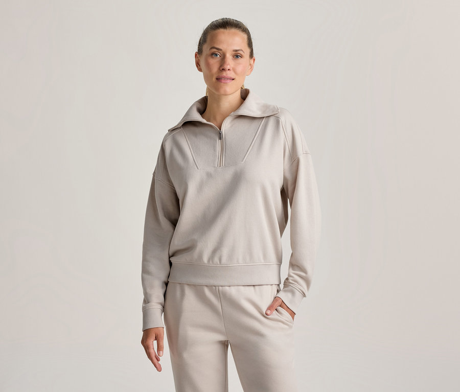 Frau steht, trägt ein hellbeiges Half-Zip-Sweatshirt und passende Jogginghose, eine Hand in der Tasche, blickt nach vorne.