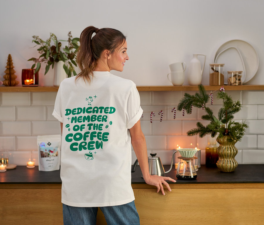 Eine Frau steht in einer Küche und trägt ein T-Shirt mit der Aufschrift "Dedicated Member of the Coffee Crew".