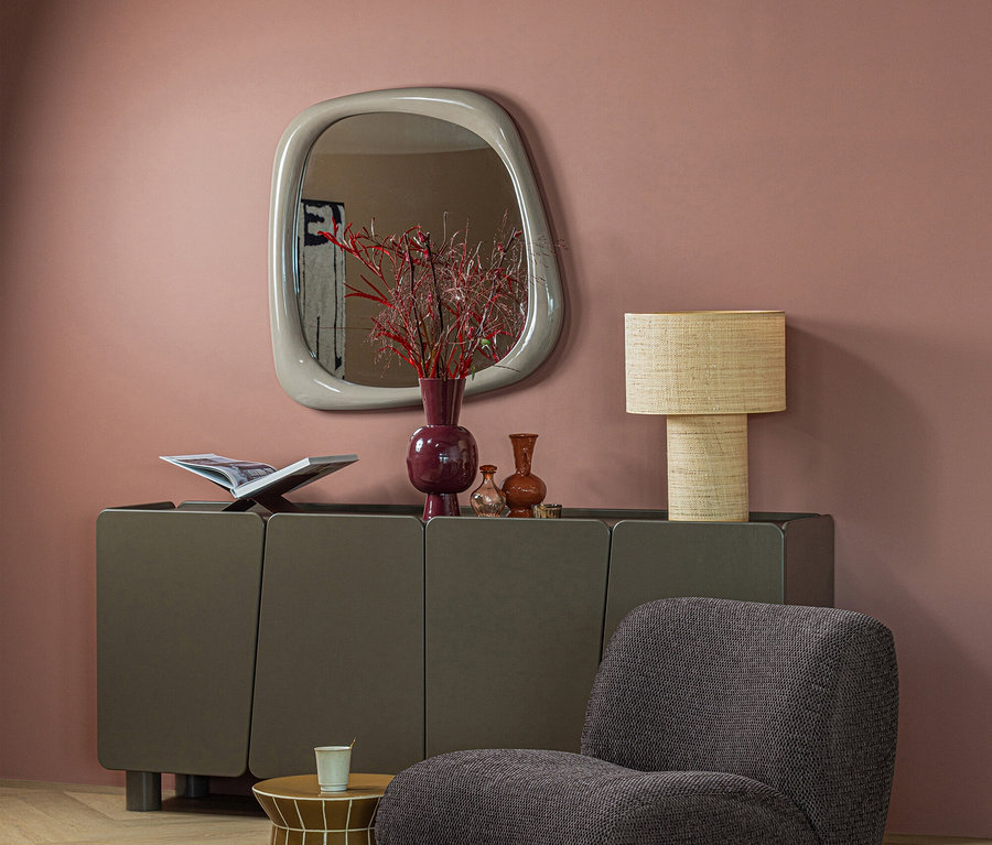 Modernes Zimmer mit altrosafarbener Wand, dunkelgrauem Sideboard, grauem Sessel und goldenem Beistelltisch auf gemustertem Teppich. Ein Spiegel, rote Vase und geflochtene Lampe schmücken das Sideboard.