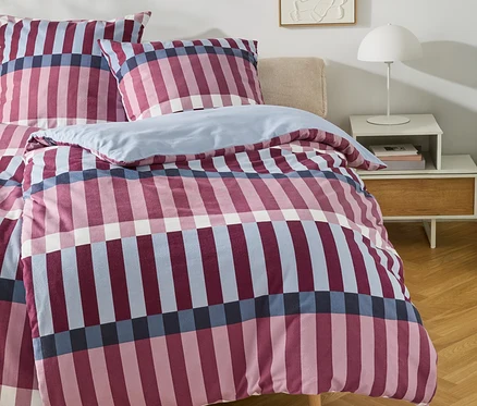 Ein Bett mit einem karierten Bettbezug in Rosa, Marineblau und Hellblau, dazu passende Kissen. Rechts daneben steht ein weißer Nachttisch mit einer weißen Lampe und Büchern. An der Wand hängt ein abstraktes weißes Reliefbild.