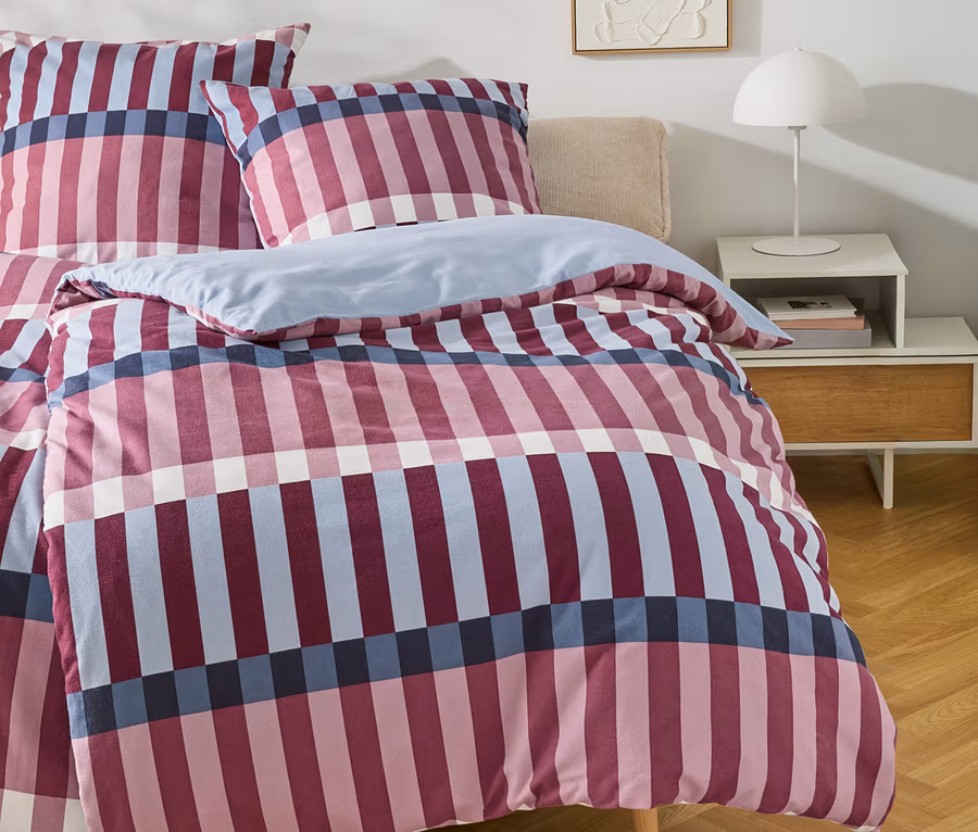 Ein Bett mit einem karierten Bettbezug in Rosa, Marineblau und Hellblau, dazu passende Kissen. Rechts daneben steht ein weißer Nachttisch mit einer weißen Lampe und Büchern. An der Wand hängt ein abstraktes weißes Reliefbild.