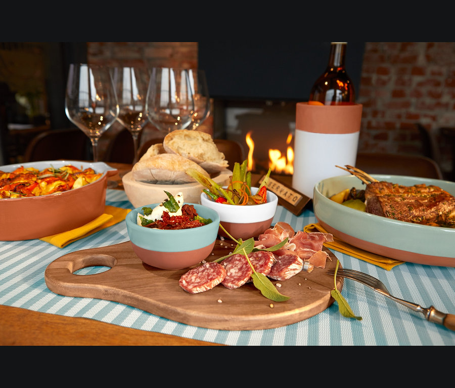 Auf einem blau-weiß gestreiften Tischläufer stehen ein Servierbrett mit Salami, Schinken und zwei kleinen Schälchen, eine Servierschale, eine Terrakotta-Ofenform, ein Tischläufer und ein Flaschenkühler.