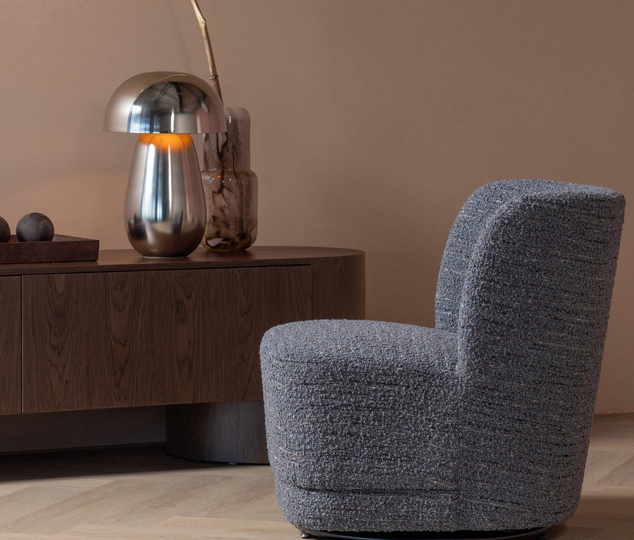 Grauer, texturierter Sessel in einem modernen Wohnzimmer. Dahinter steht ein dunkler Holzschrank mit einer silbernen Pilzlampe, einer rauchigen Vase mit Ast und Deko-Kugeln. Warme braune Wand, heller Holzfußboden.