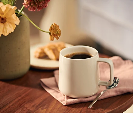 Eine hellbeige Kaffeetasse mit schwarzem Kaffee steht auf einer rosa Serviette auf einem Holztisch, neben einer Vase mit orangefarbenen und rosa Blumen und einem Teller mit Gebäck.